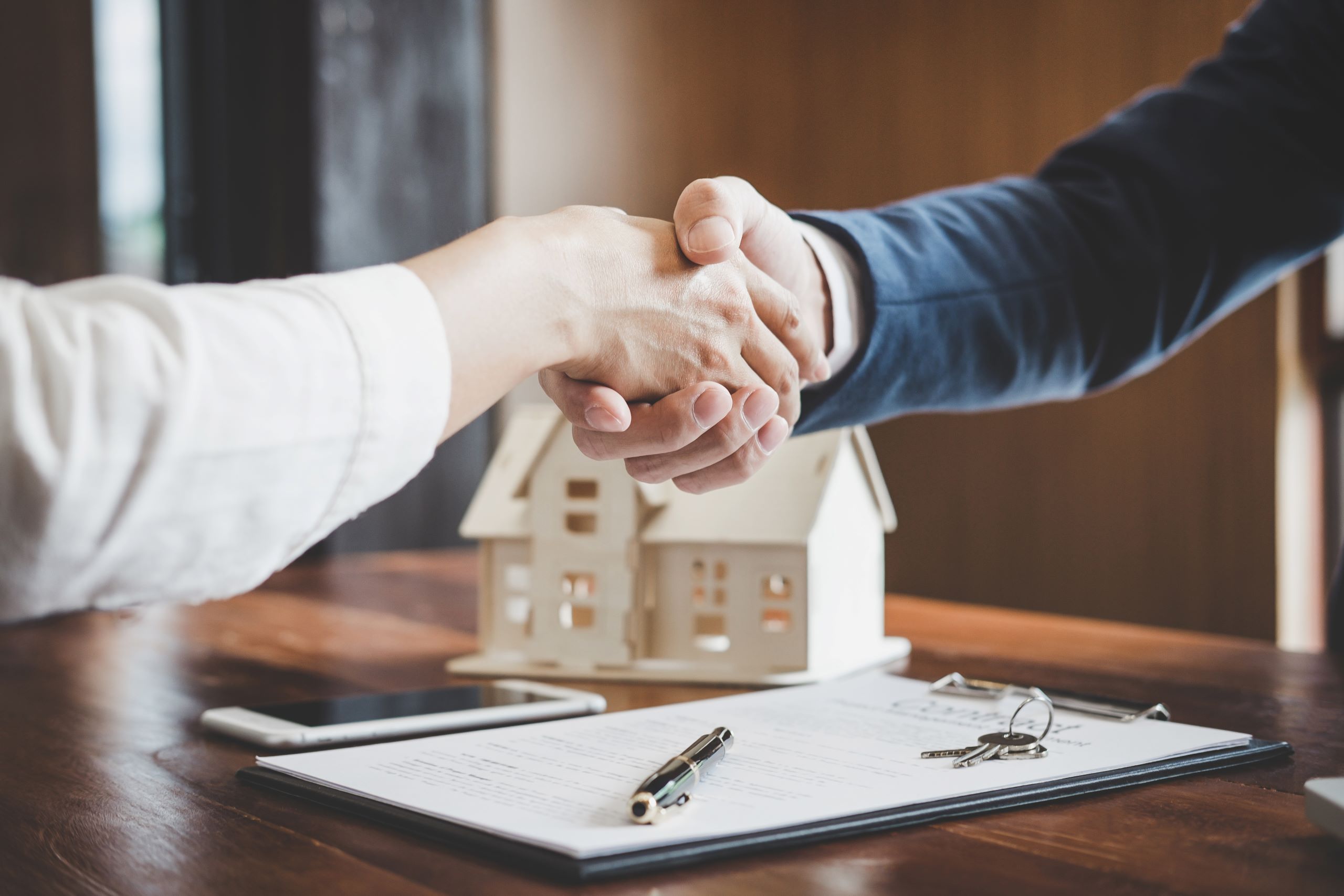 Two individuals shaking hands over a real estate contract, with a miniature house model and a set of keys on the table, symbolizing a property agreement.