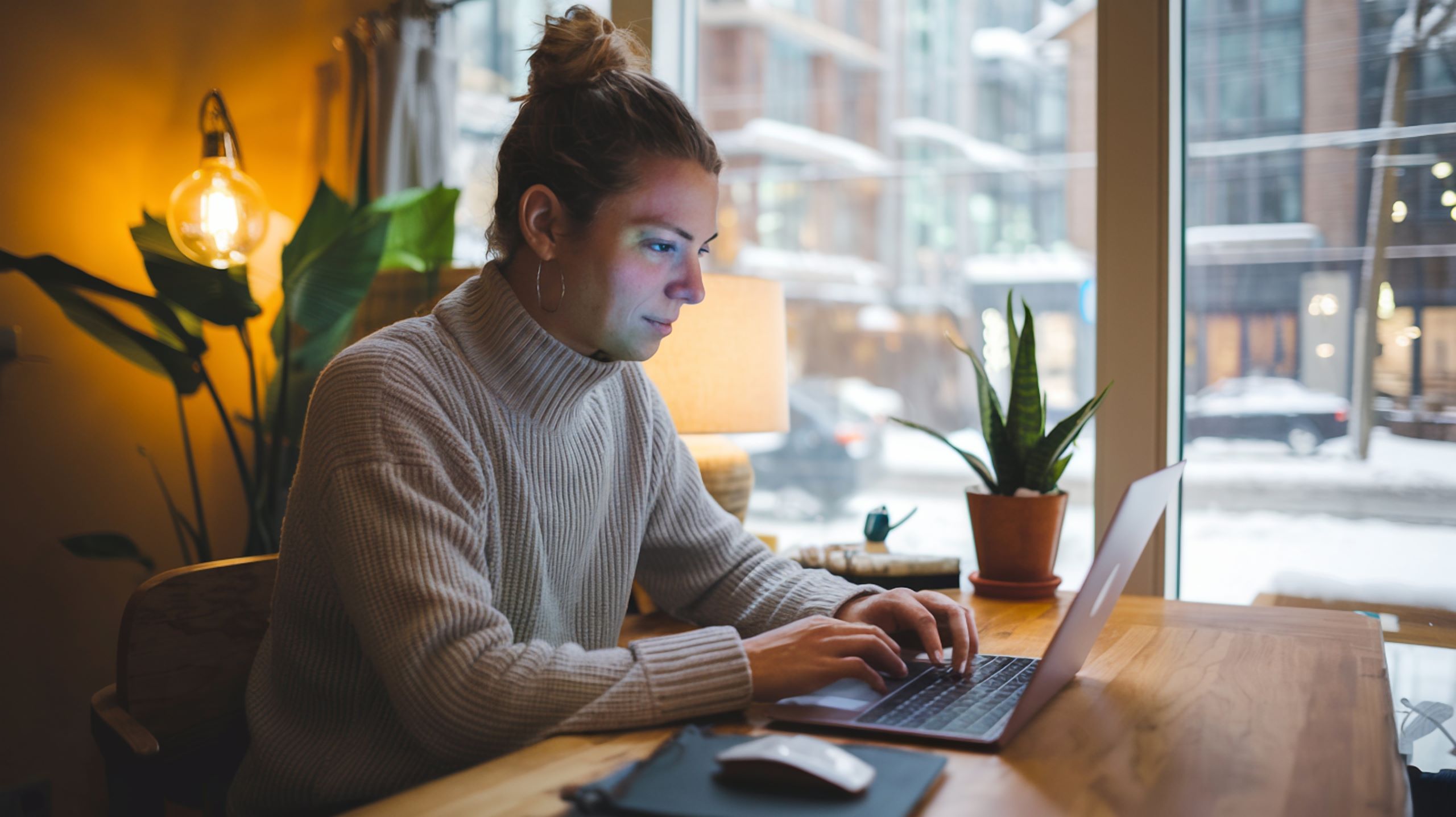 Person working on a laptop in a cozy café setting with warm lighting, plants, and a snowy cityscape visible through large windows.